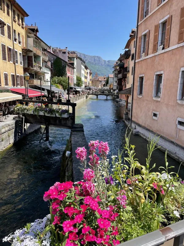 A canal in Annecy’s old town lined with pastel buildings, café terraces and pink flowers, with mountains in the distance