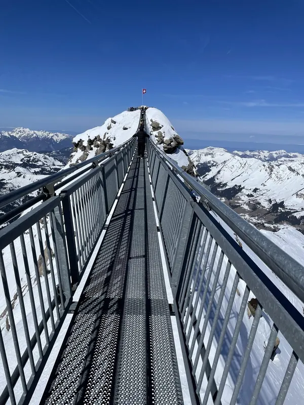 Looking along the Peak Walk suspension bridge towards a snowy summit with a Swiss flag