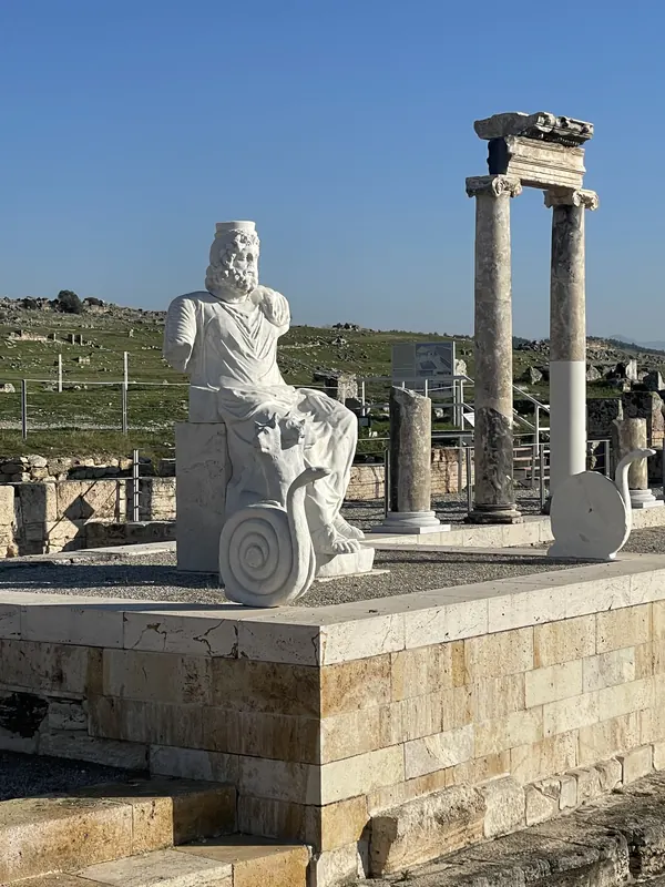 A white marble statue of a seated figure beside standing columns in ancient Hierapolis