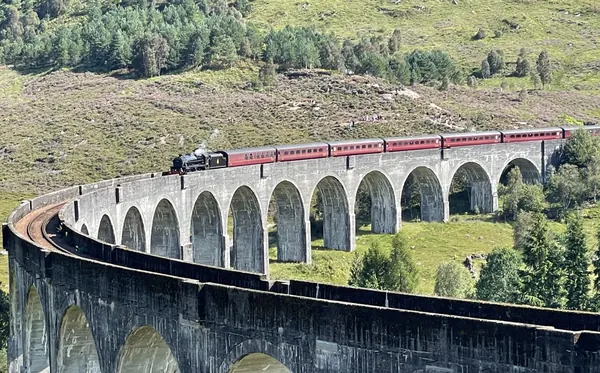A steam locomotive pulling red carriages across the curving stone arches of the Glenfinnan Viaduct