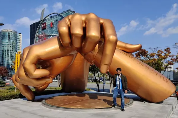 Saptarshi with a conference badge standing by the giant golden Gangnam Style hands statue