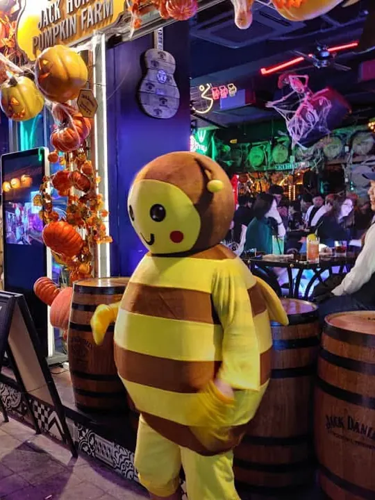 A cheerful bee mascot outside a bar decorated with pumpkins and cobwebs for Halloween