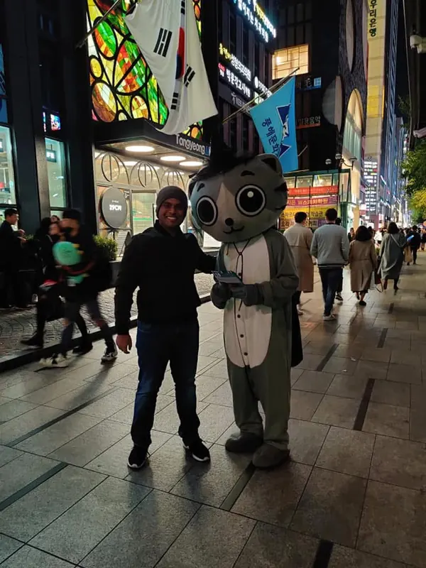 Saptarshi smiling beside a large grey cat mascot on a brightly lit Seoul street at night